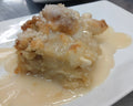 A plated dessert serving with a golden-brown crumb topping and creamy light brown sauce on a white square plate, with another plated dessert blurred in the background on a stainless steel surface.