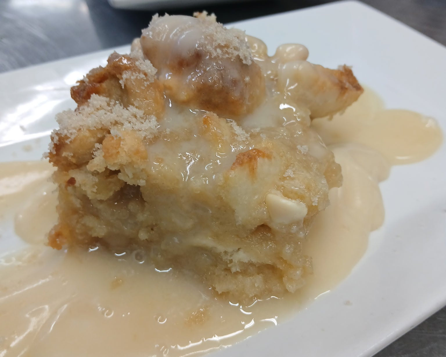 A plated dessert serving with a golden-brown crumb topping and creamy light brown sauce on a white square plate, with another plated dessert blurred in the background on a stainless steel surface.