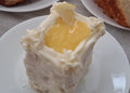A square slice of lemon cake with white frosting and a glossy yellow lemon filling on a white plate.